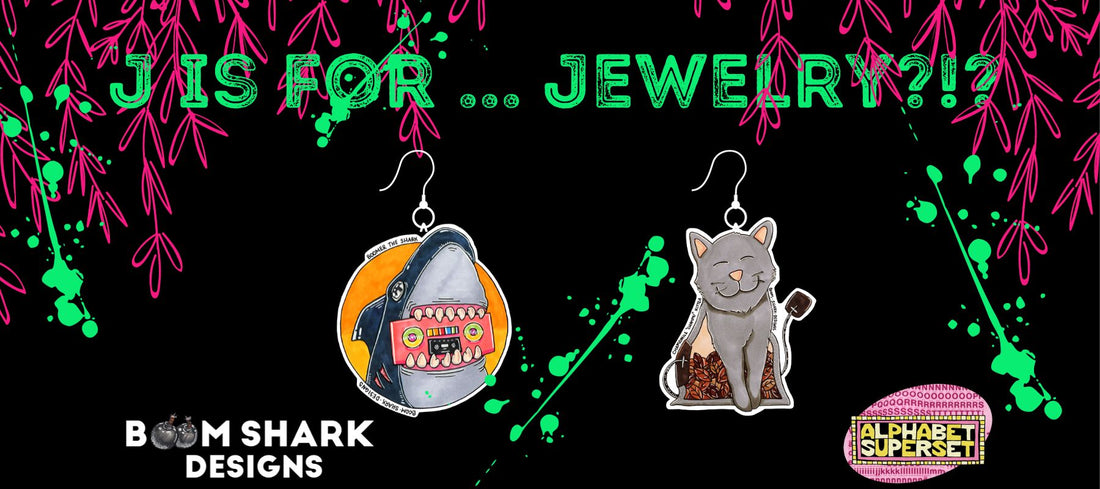 on a black background the text reads "J is for Jewelry?" and includes an earring of boomer the shark and an earring of KitTea the Tea Cat with the image tags for boom shark designs and the alphabet superset.